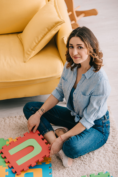 overhead view of smiling woman in jeans sitting on carpet with puzzle mat