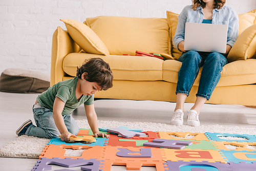 cute child in green t-shirt playing with puzzle mat while mother using laptop