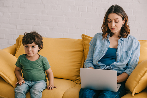 Cute kid sitting on sofa while mother using laptop