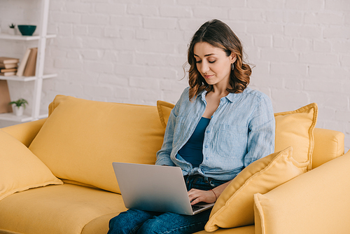 Attractive freelancer sitting on yellow sofa and using laptop