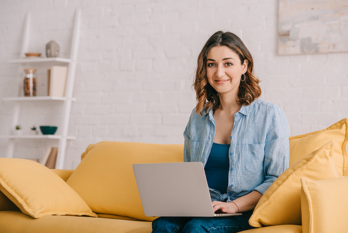 Attractive smiling freelancer sitting on yellow sofa and using laptop