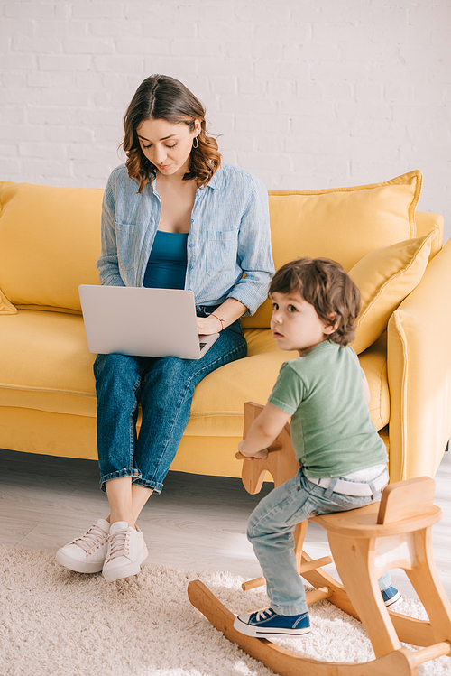 child sitting on rocking horse while mother working with laptop in living room