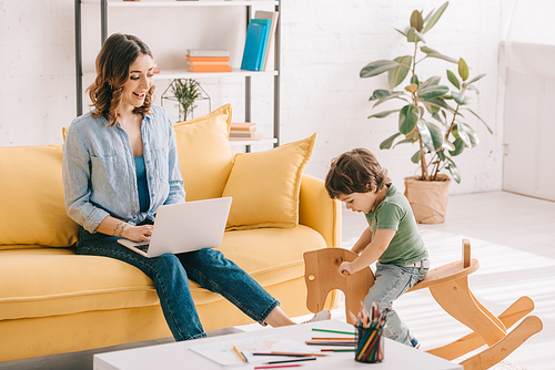 kid sitting on rocking horse while mother working with laptop in living room