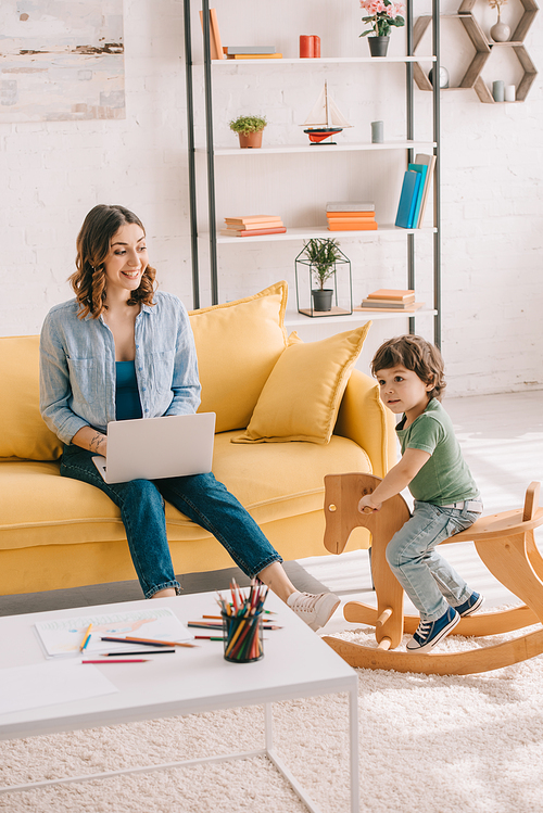 kid sitting on rocking horse while mother working with laptop in living room