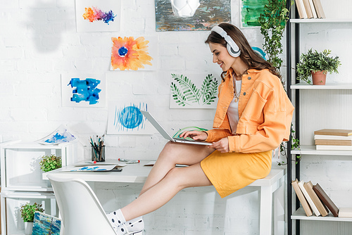 smiling trendy woman in headphones using laptop while sitting on desk