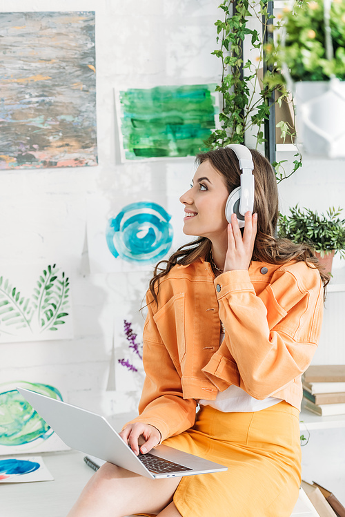 happy woman looking at paintings on wall while listening music in headphones and using laptop