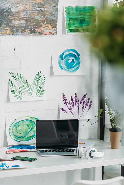 selective focus of laptop with blank screen, headphones and paper cup on tabletop near paintings