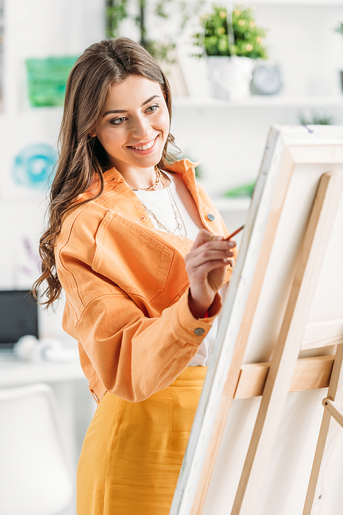 cheerful young artist in trendy clothing painting on easel at home