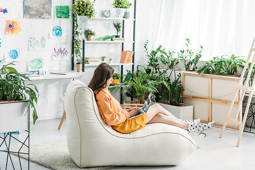 young woman using laptop while sitting on soft chaise lounge in light spacious room