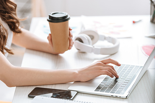 cropped view of young woman holding paper cup while using laptop near headphones