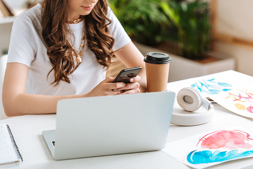 cropped shot of young woman using smartphone while sitting near laptop and disposable cup