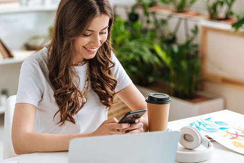 happy beautiful woman using laptop while sitting at desk near laptop and paper cup