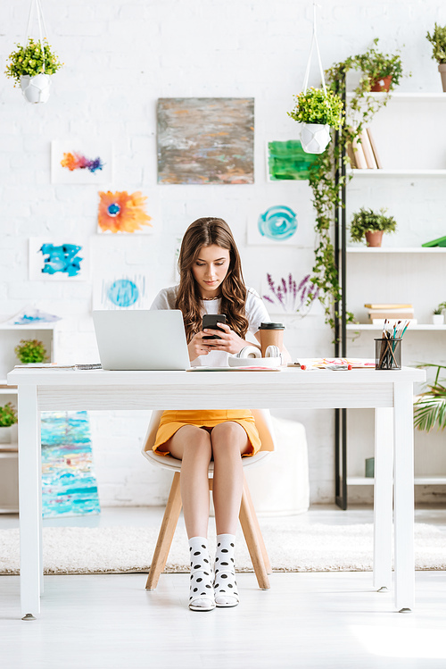 young woman using smartphone while sitting at desk in spacious room with paintings on wall