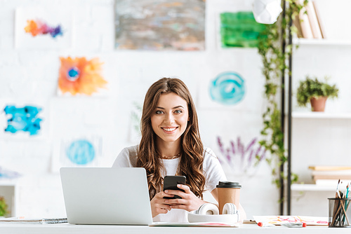 cheerful woman  while sitting at desk near laptop and using smartphone