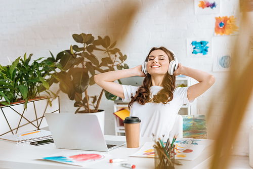 selective focus of happy girl in headphones stretching while sitting at desk near laptop, disposable cup and paintings