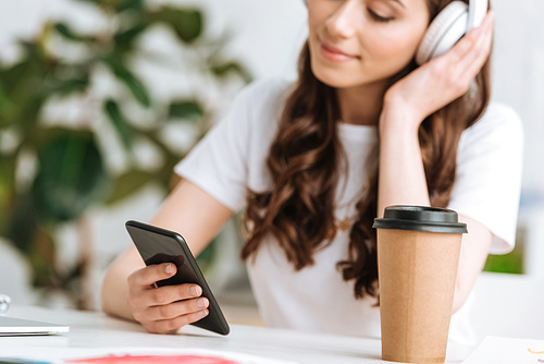 selective focus of young woman in headphones using smartphone while sitting near disposable cup