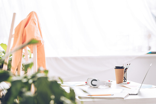 selective focus of desk with paper cup and headphones near window with white curtain