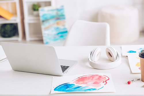 laptop, headphones and paintings on white tabletop near colorful paitings