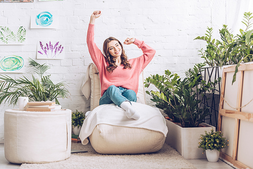 smiling girl stretching while sitting on soft chaise lounge near pouf and lush green plants