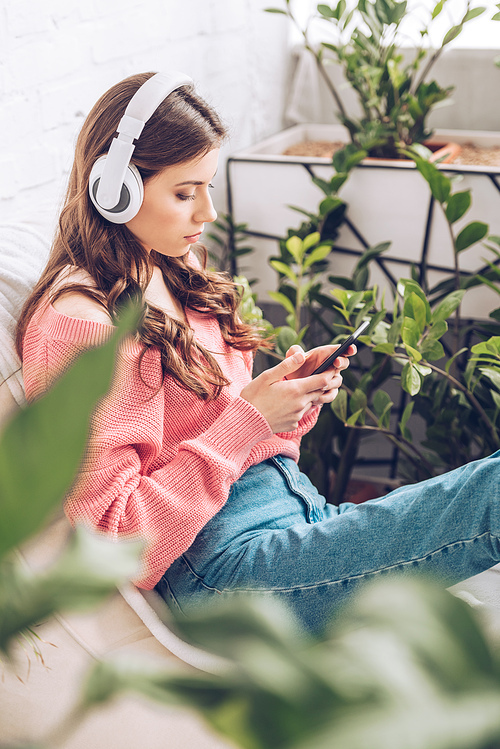 selective focus of pensive girl listening music in headphones and using smartphone while sitting surrounded by green plants