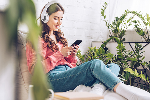selective focus of smiling girl using smartphone while sitting surrounded by green plants and listening music in headphones