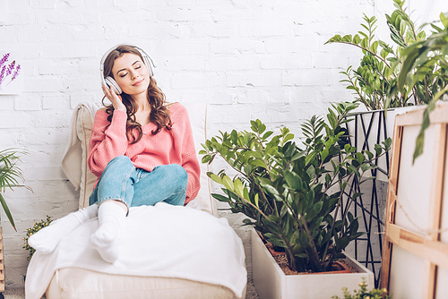 pretty girl with closed eyes relaxing while sitting in soft chaise lounge and listening music in headphones