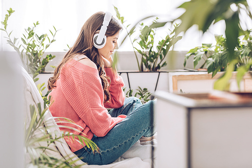 pensive young woman listening music in headphones while sitting surrounded by green plants at home