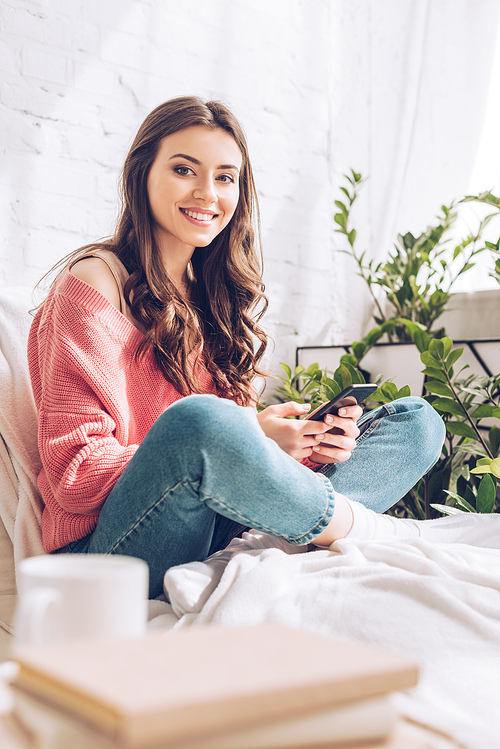 selective focus of beautiful girl smiling at camera and using smartphone while sitting with crossed legs at home