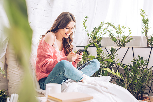 selective focus of cheerful girl using smartphone while sitting with crossed legs near green plants at home