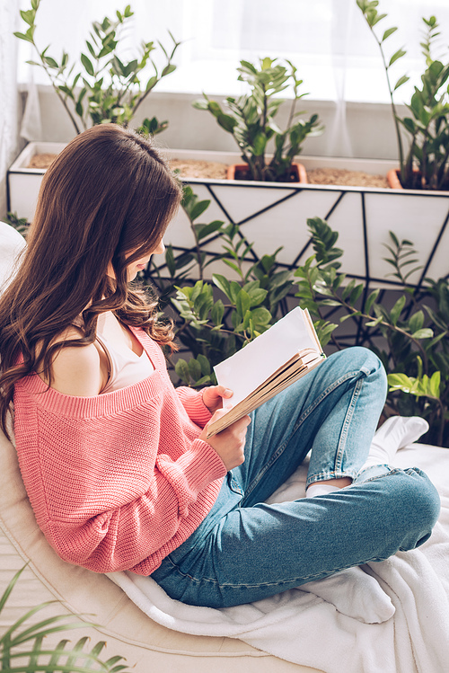 young woman reading book while sitting surrounded by green plants at home