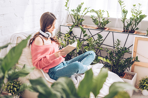 selective focus of young woman reading book while sitting with crossed legs surrounded by lush plants at home