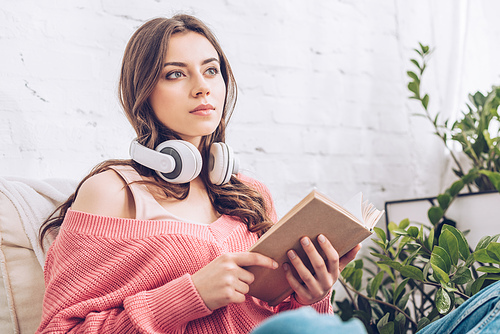 pensive woman with headphones on neck holding book and looking away