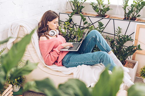 selective focus of pretty girl with headphones on neck using laptop while sitting on soft chaise lounge