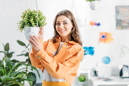 cheerful woman touching hanging flowerpot with green plant and smiling at camera