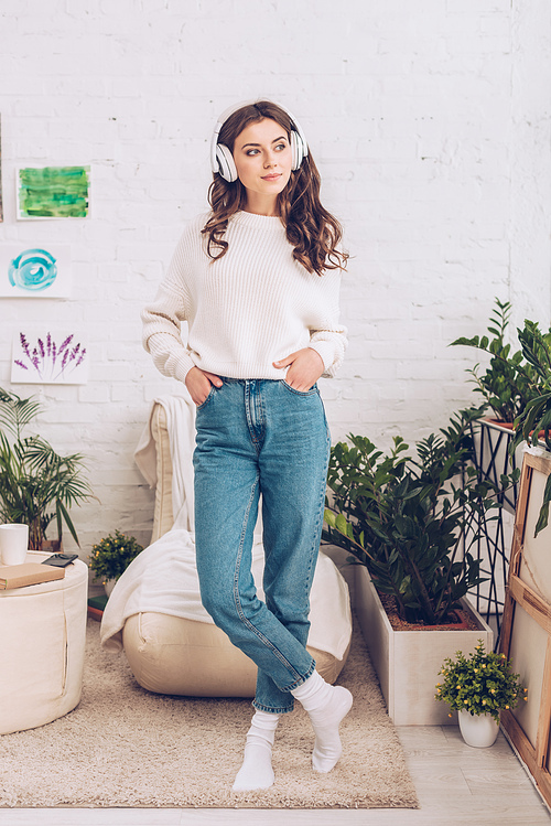 smiling attractive girl listening music in headphones while standing with hands in pockets and looking away