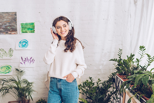 cheerful girl listening music in headphones while holding hand in pocket and looking away