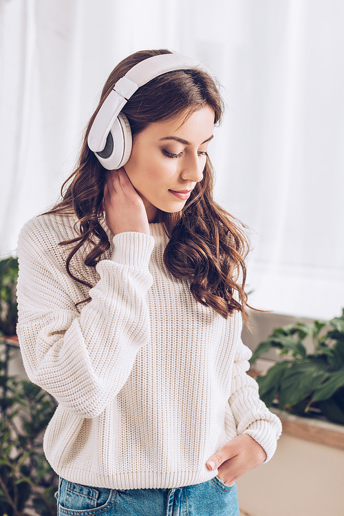 pensive young girl listening music in headphones, holding hand in pocket and looking down