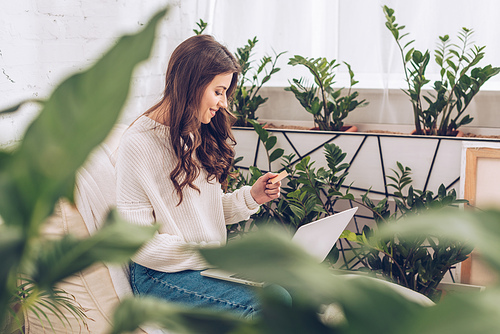 selective focus of smiling woman holding credit card and using laptop while sitting surrounded by green plants  at home