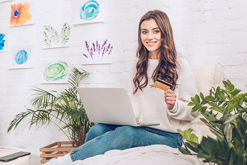 happy girl using smartphone and holding credit card while smiling at camera