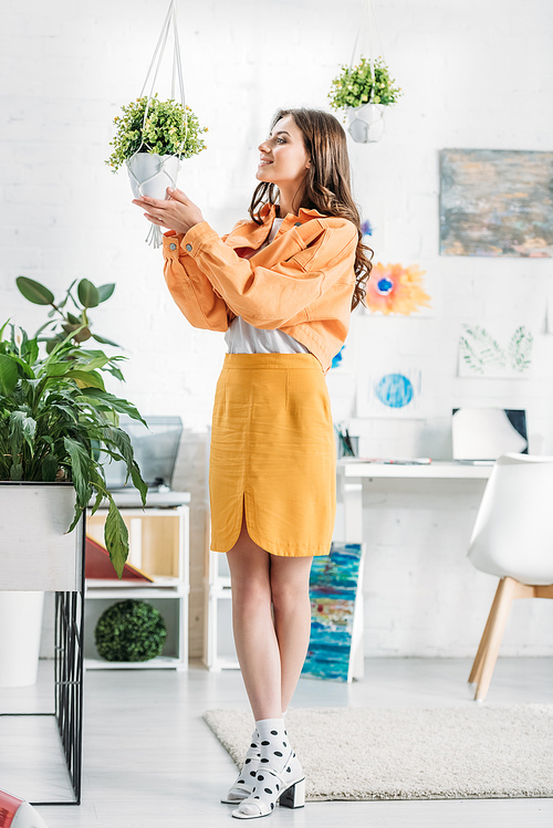 trendy woman in orange clothing touching flowerpot with green plant
