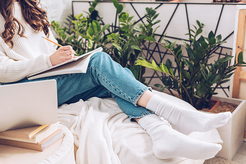 cropped view of young woman writing in notebook while sitting near green plants at home