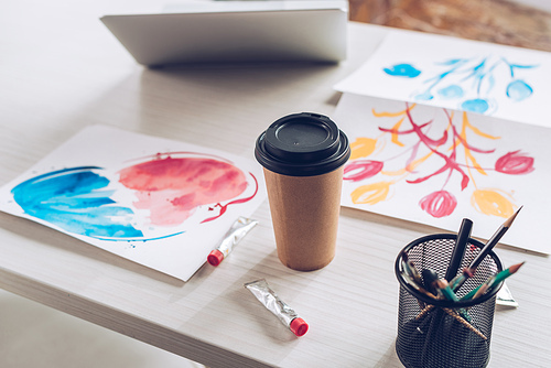 tabletop with laptop, disposable cup, paint tubes, stationery and colorful paintings