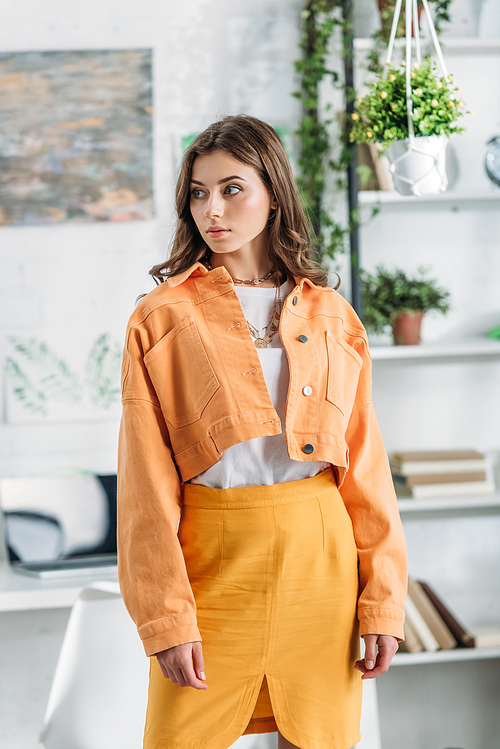 pensive pretty girl in orange clothing looking away while standing near rack with books and plants