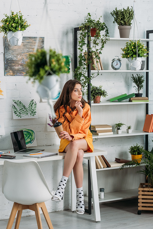 pensive young woman sitting on desk in room decorated with green plants and paintings on wall