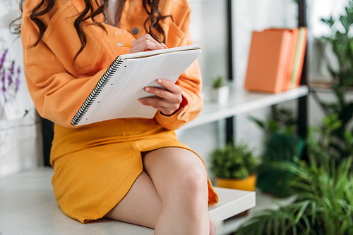 partial view of young woman sitting on desk and writing in notebook