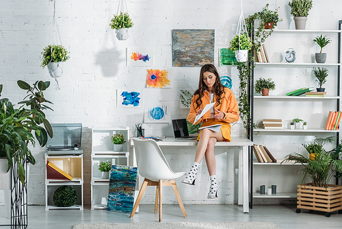 thoughtful young woman sitting on desk in light spacious room decorated with green plants and paintings on wall