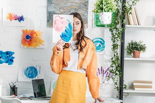 beautiful girl hiding face behind painting while standing in spacious room decorated with green plants and drawings on white wall