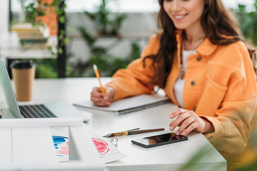 partial view of smiling girl sitting at desk, using smartphone and writing in notebook