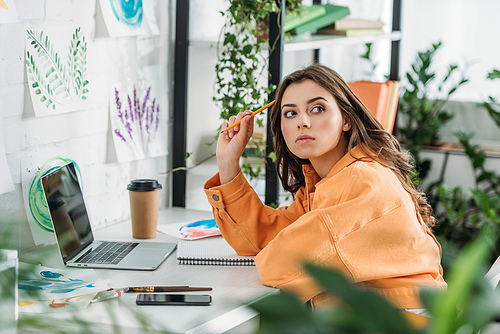 selective focus of thoughtful young woman sitting at desk near laptop and looking away