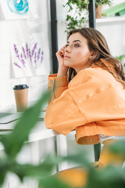 selective focus of pensive young woman in orange closing looking away while sitting at desk at home